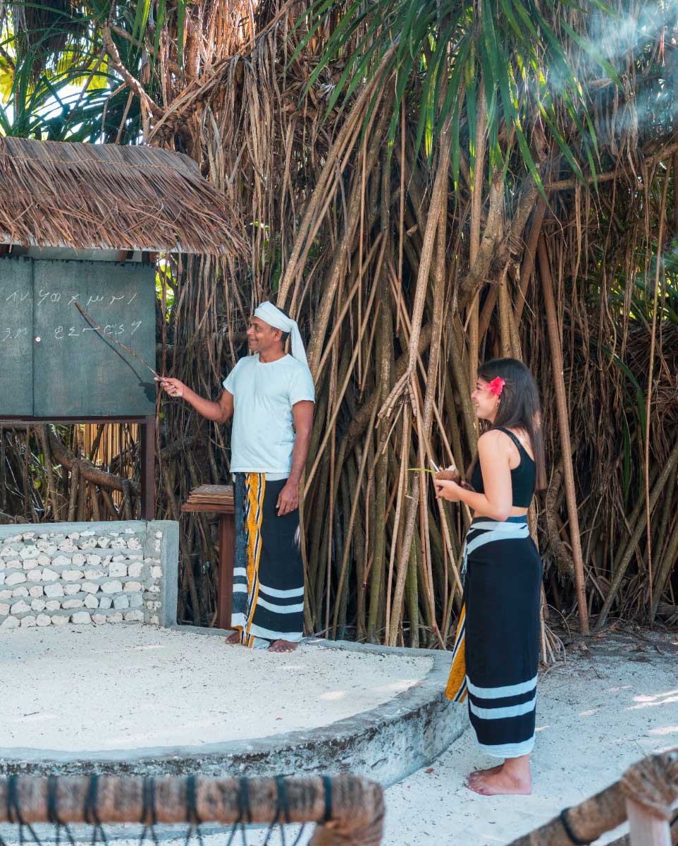 A teacher points to Dhivehi words written in Thaana script on a blackboard while a guest looks on at Heritance Aarah